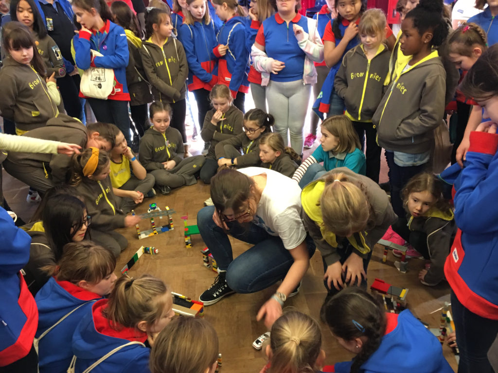 Guides, brownies and rangers gathered round different model bridges made of Lego. A woman sits in the middle of the group testing the bridges with a toy car